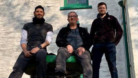 Aron Sapsford, a middle-aged man wearing glasses with brown hair sitting in the middle of two younger men, in front of the white farmhouse building.