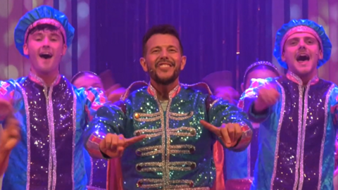 Three people in a pantomime on stage. They are singing and smiling. A man with short dark hair in the centre is wearing a green sparkly coat with gold buttons. The two people either side of him are wearing blue sparkly coats with purple stripes running vertically down the centre