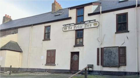 The front of the boarded up La Folie Inn pub in Jersey. There is graffiti on the windows and the building is in a general state of disrepair. 