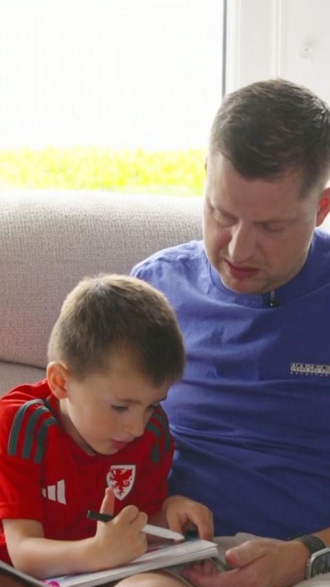 A man in a blue t-shirt, and a young boy with a red Wales football t-shirt are looking down at a coloring book. 