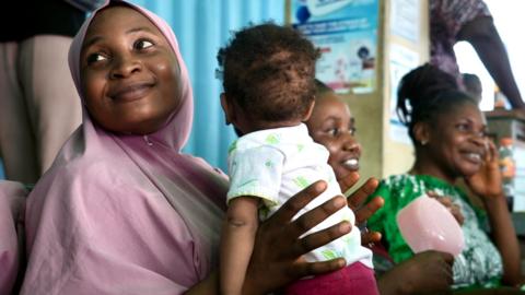 A woman in a pink headscarf smiles while she holds a small child in front of her.