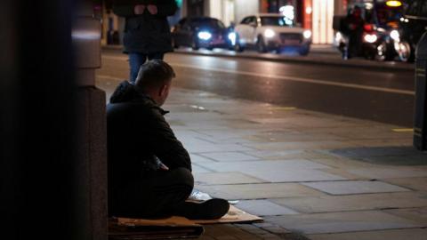 A man sits on cardbard in a doorway on the pavement.  It is dark and cars with their lights on drive by, and a person on the pavement is walking past.