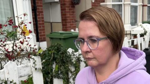 Jenefer Sully, wearing a pink hoodie, has glasses and short brown hair that is swept over to one side. Behind her are red brick terraced houses with front gardens.