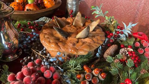 A pastry pie sits in the middle of a Christmas table, surrounded by nuts, fruits and foliage