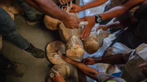 The image shows a food distribution scene where several people are handing out or receiving bags of bread. Multiple hands are visible reaching for or holding plastic bags containing round loaves.