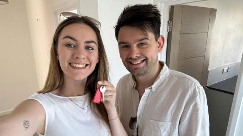 Krista has brunette hair and is wearing a white top. She is holding a key with a red keyring. Mikey is standing to her right and is wearing a light grey shirt. He has short dark brown hair and short stubble on his face.