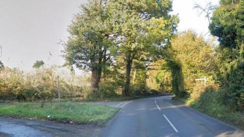 Weston Hall Road bends to the right, while there is a triangle junction off to the left and give way markings. The road is bordered by trees, hedging and grass.