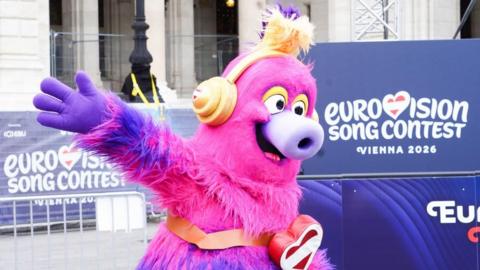 The 2026 Eurovision Song Contest's official mascot, Auri, who is furry and bright pink and purple and wears yellow headphones, smiles and waves next to a Eurovision Song Contest sign