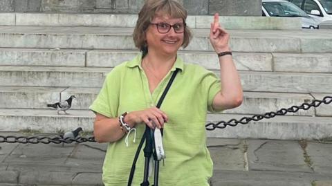 Jules wearing a green blouse, and glasses with walking sticks in her hand. She is smiling at the camera and pointing up.