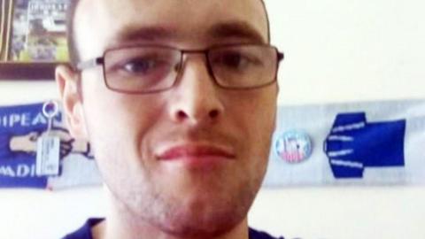 A man with short brown hair and glasses looks at the camera. He appears to be in a bedroom or living room