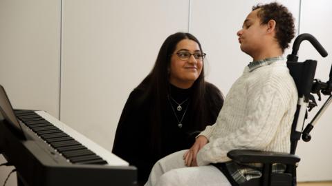 Karima Motala sits beside her son Jaden, who is in a wheelchair next to a keyboard, looking towards him in a bright room.