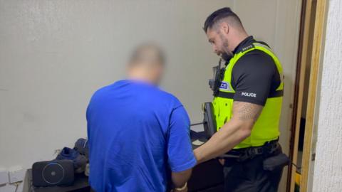 A police officer wearing a yellow vest and black shirt hold the wrist of a man with a blue shirt and a blur added to his head