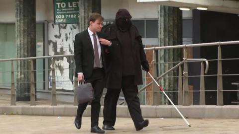 Sean Pullen, who wears a hat, a black face covering and dark glasses, walks towards the court building with a white case extended in front of him and linking arms with a younger man in a black suit carrying a laptop bag 