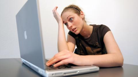 A woman is looking at her grey laptop with an animatedly confused expression on her face. She has blonde hair which is held back by a head band and is wearing a brown t-shirt.