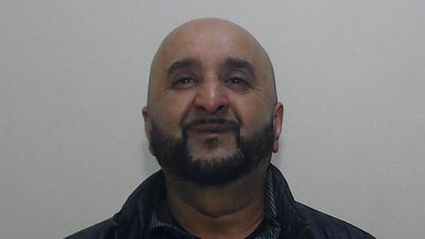 Police custody shot of Ajaz Ahmed who is bald with a black beard wearing a black jacket and black shirt