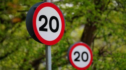Two round 20mph speed limit signs against a backdrop of green trees - one sign is in the foreground and one is further away and out of focus.