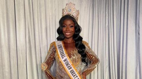A woman wearing a large gold crown and a golden dress with a yellow and white sash which says "Universal Woman Great Britain". She is standing and smiling, with her hand on her hip, in front of a white curtain.