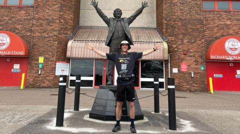 A man in a black t-shirt and black shorts stands with arms outstretched in front of a statue of a man with his arms outstretched. The man and the statue are in front of Aberdeen FC's football stadium's Richard Donald stand.
