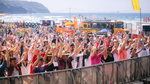 Hundreds of people stand at the barrier of a music festival on a beach with their hands in the air. It is a sunny day and you can see the sea in the background.