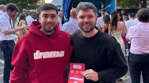 Two men standing outside and smiling, holding a poster advertising the fan zone. It reads: "Victory at the Baths".