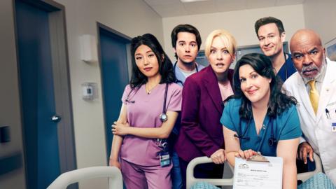 The cast of St Denis Medical, an array of Doctors leaning over the end of a hospital bed. The woman at the front is holding a clipboard and paper for the viewer