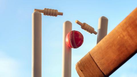 A cricket ball is hitting a wicket and the two bails are in the air in the process of being broken. A cricket bat can be seen in the bottom right corner. The blue sky behind the wicket.