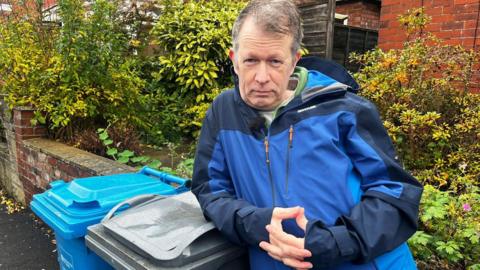 Stuart Atkinson has short grey hair and is wearing a blue waterproof jacket. He is leaning against a grey wheelie bin while looking at the camera.
© BBC