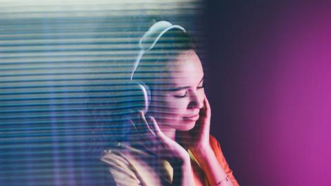 A woman with curly hair listens to music with white headphones in front of a purple background.