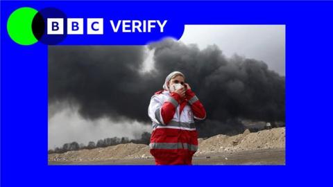 An emergency worker in Tehran shields her face as black smoke rises behind her. She is wearing a red and white jacket and a face mask and standing in a sandy location. The imagen is imposed over the BBC Verify colours and branding. 