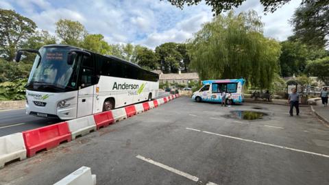 A large white Anderson Travel coach is parked on the left side of a narrow village road, opposite an ice cream van. Red and white plastic barriers line the roadside. A few pedestrians walk nearby.