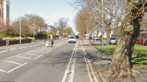 A Google street view shot of New Chester Road, near the junction with Platt Grove in New Ferry, Birkenhead, showing an empty, urban main road with a car in the distance and a tree to the right.