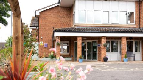 Amberley Hall Care 鶹Լ in King's Lynn is a two-storey building with large white framed windows and doors surrounding the ground floor reception. Blue pots filled with flowers stand alongside the brick pillars. 