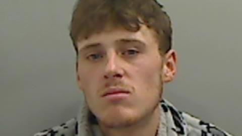 A blurry mugshot of Bradley Thomas who has short, brown hair and stubble on his upper lip and chin. His lips are pursed and he is wearing a grey and black patterned top.