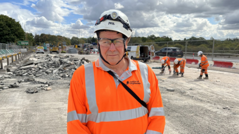 A head and shoulders shot of Terry Robinson. He is wearing a fluorescent orange shirt with a collar. He is also wearing safety glasses and a hard hat with a torch on the front. Behind him is the over bridge on the A40. Three workers are immediately to his right in the background hitting and drilling the concrete on the bridge. There's a white van with its double doors open over Robinson's right shoulder with a car passing through the single lane of traffic set up on the other side. To his left is a large pile of broken up concrete rubble. In the background there are various other construction vehicles, a green tree line behind that and a cloudy sky streaked with some blue spots.