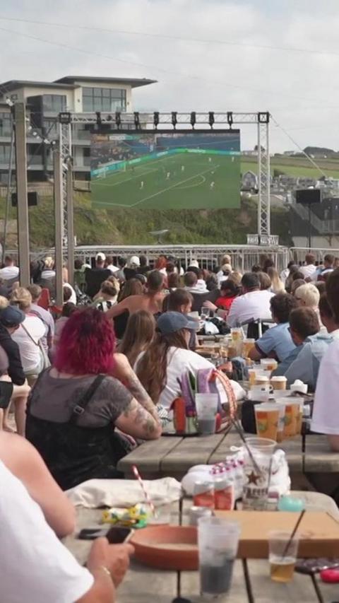 A crowd of people sitting at tables on a beach watching a big screen showing football.