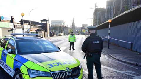 A yellow, blue and black Skoda police car parked with an officer in a green uniform standing next to it. A police officer is standing behind the car in a hi-vis police jacket, he is standing in front of police tape which is across the road.