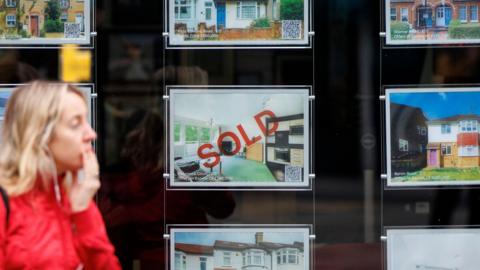 An estate agent's window showing a house being advertised says 'SOLD' in red across it.