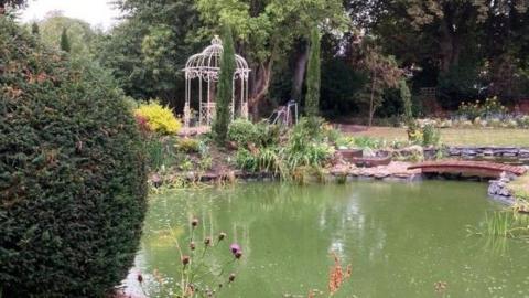 A large pond is surrounded by planting and a white obelisque structure. A little bridge crosses the stream and purple thistles are in the foreground.