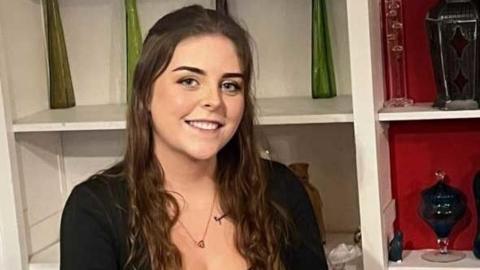 A young woman with long brown hair, wearing a black long sleeved top and a necklace. Behind her are white and red shelves with green and black pottery on them.