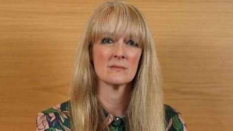 Nicola Killean has a serious expression. She has long, blonde hair with a fringe and is wearing a green floral top.