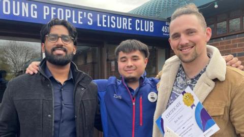 A man with dark hair, a beard and a black jacket and a man with blonde hair and a brown jacket on either side of a man with dark hair and a blue Tesco top