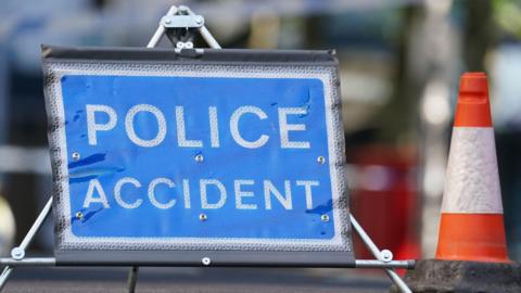 A blue road sign that reads "police" with the word "accident" underneath but in white writing. An orange and white traffic cone sits next to it.