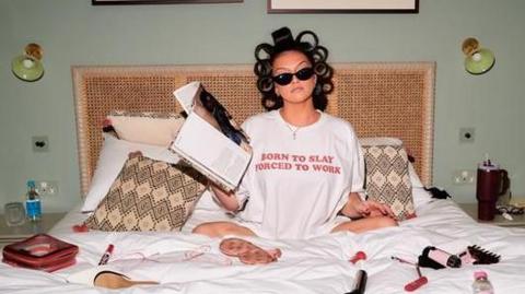 A woman with her hair in rollers and wearing sunglasses and a T-shirt reading "born to slay forced to work" sits on a double bed surrounded by makeup products, hair styling tools and shoes. She is holding a magazine.