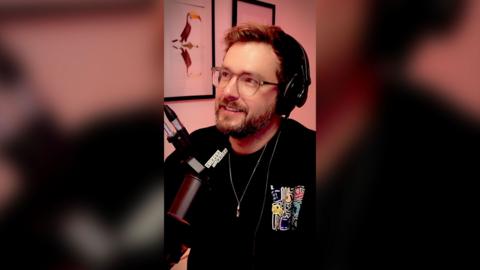 Iain Stirling wears black headphones and smiles. He is wearing a black jumper and glasses.