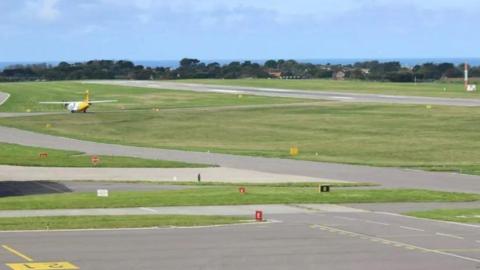 Guernsey airport runway. Grass is visible as well as grey tarmac runways. A yellow and white plane can be seen from behind in the distance. 