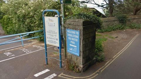 A blue entrance sign to the Shoalstone Seawater Pool with a white arrow pointing left towards a car park. The sign is on a concrete pillar.