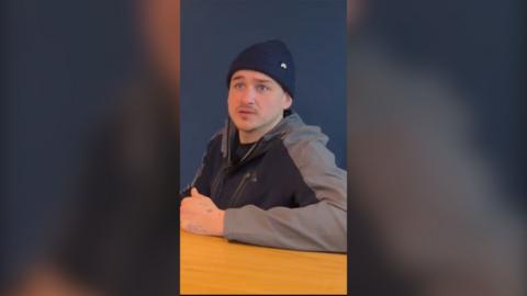 A man wearing a black and grey zip up jumper and a black beanie