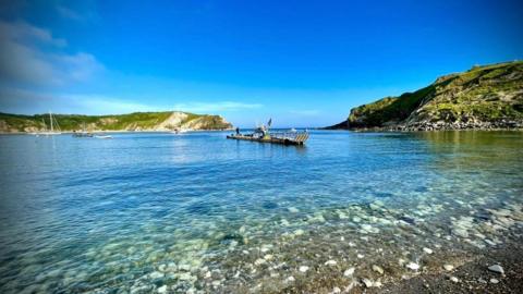 A sunny day at Lulworth Cove with clear blue water. The cover is surrounded by cliffs with green grass and you can see the narrow entrance in the distance. A pontoon is moored in the cove with three boats. 