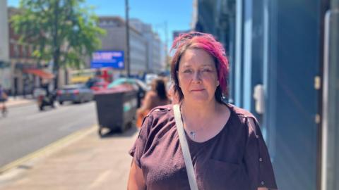 A woman with red hair stands outside a blue building. It is a sunny day.