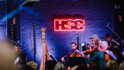 An orchestra performing on a stage with a fluorescent sign stating "HSC".